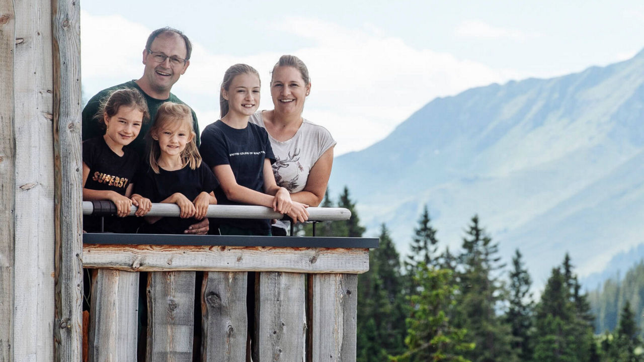 Familie steht auf dem Balkon ihrer Alm, hinter ihr die Berge