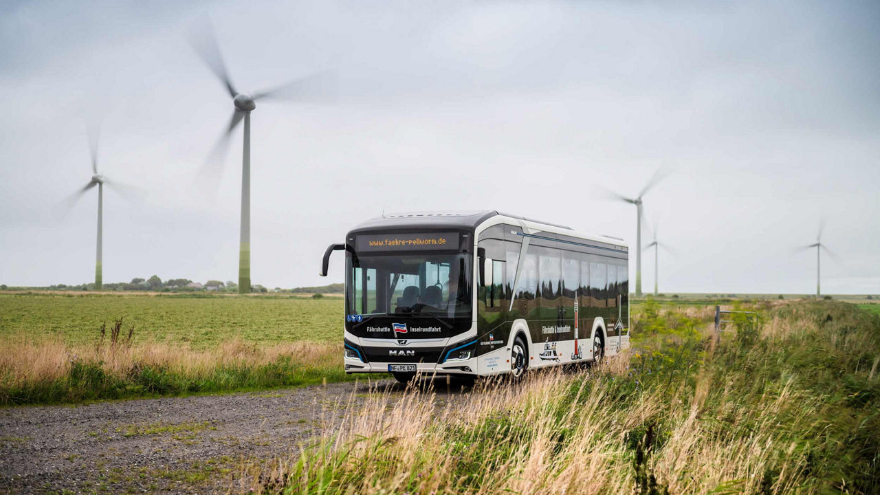 MAN Lion's City E on a green road and lots of wind turbines in the distance