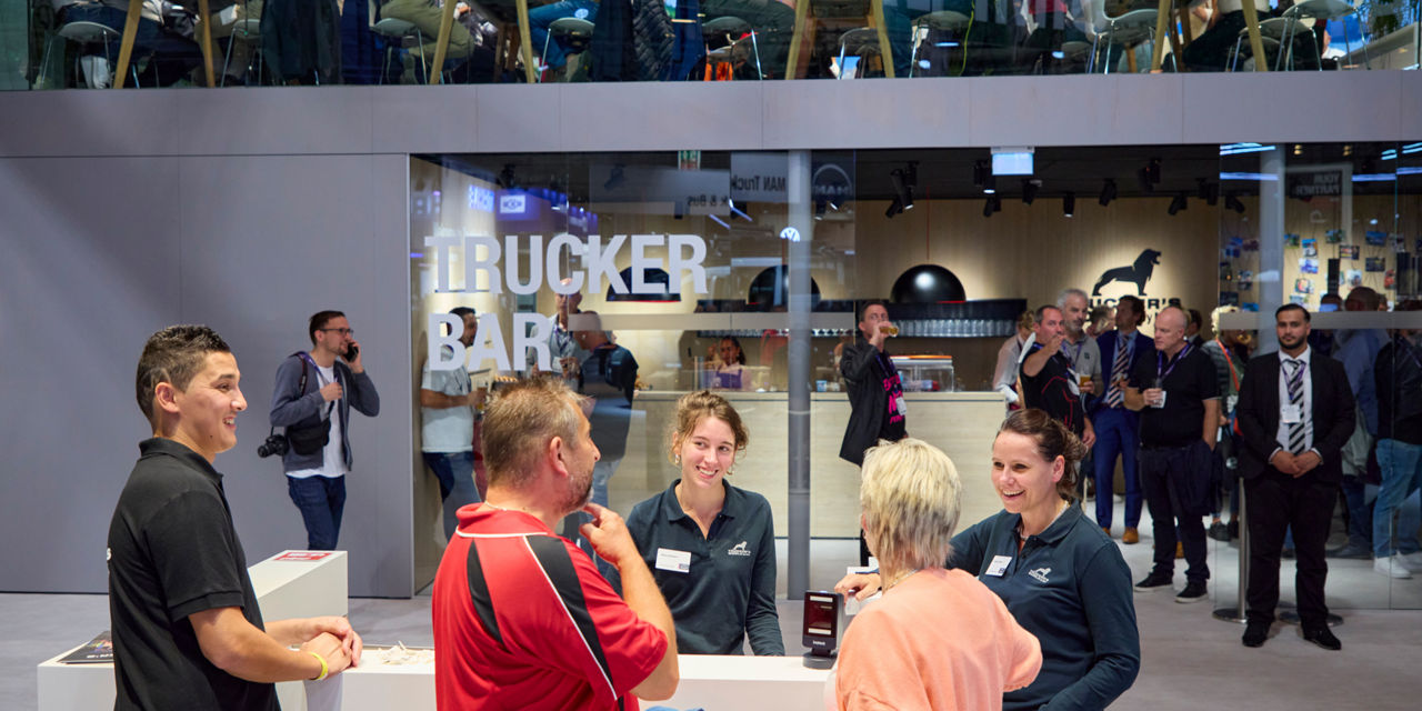 Menschen stehen an einem Stand auf einem Messegelände, im Hintergrund sieht man Menschen etwas trinken, in einem Raum, auf dessen Wand "Trucker Bar" steht.