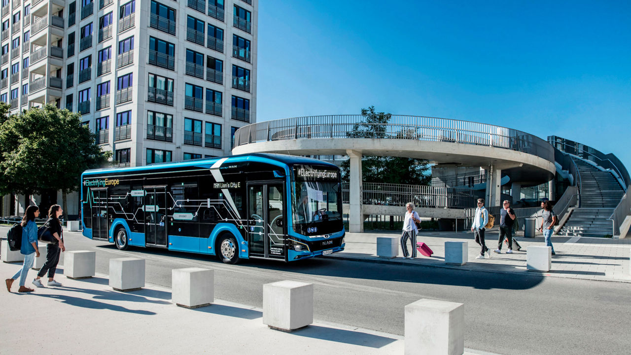 Un MAN Lion’s City E est arrêté devant un bâtiment, des passagers avec des valises montent dans le bus électrique.