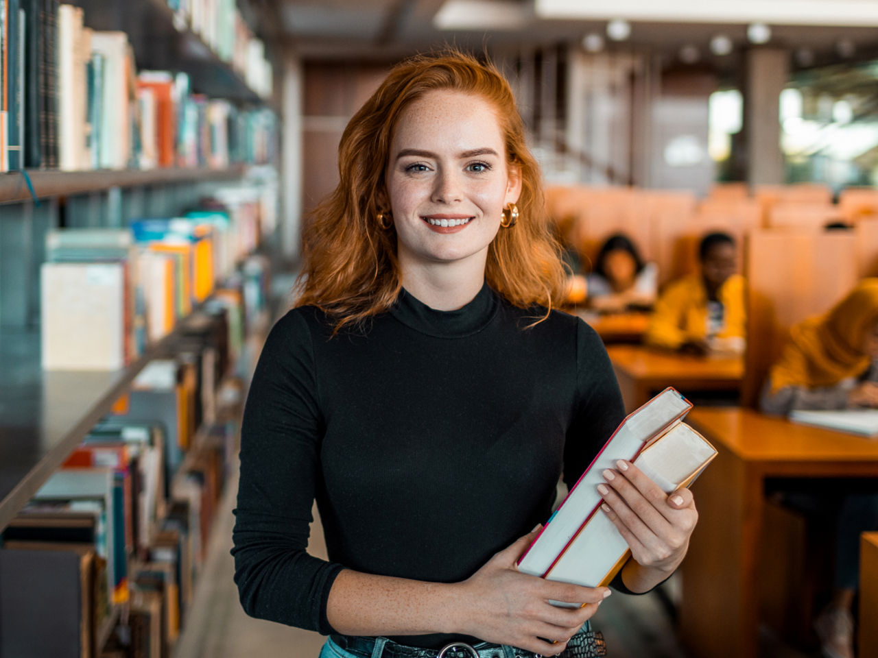 A young woman is standing in a library and is holding books in her hand