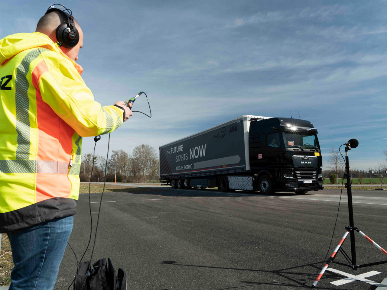 Ein Mann in Warnweste hält ein Gerät, ein Lkw fährt im Hintergrund