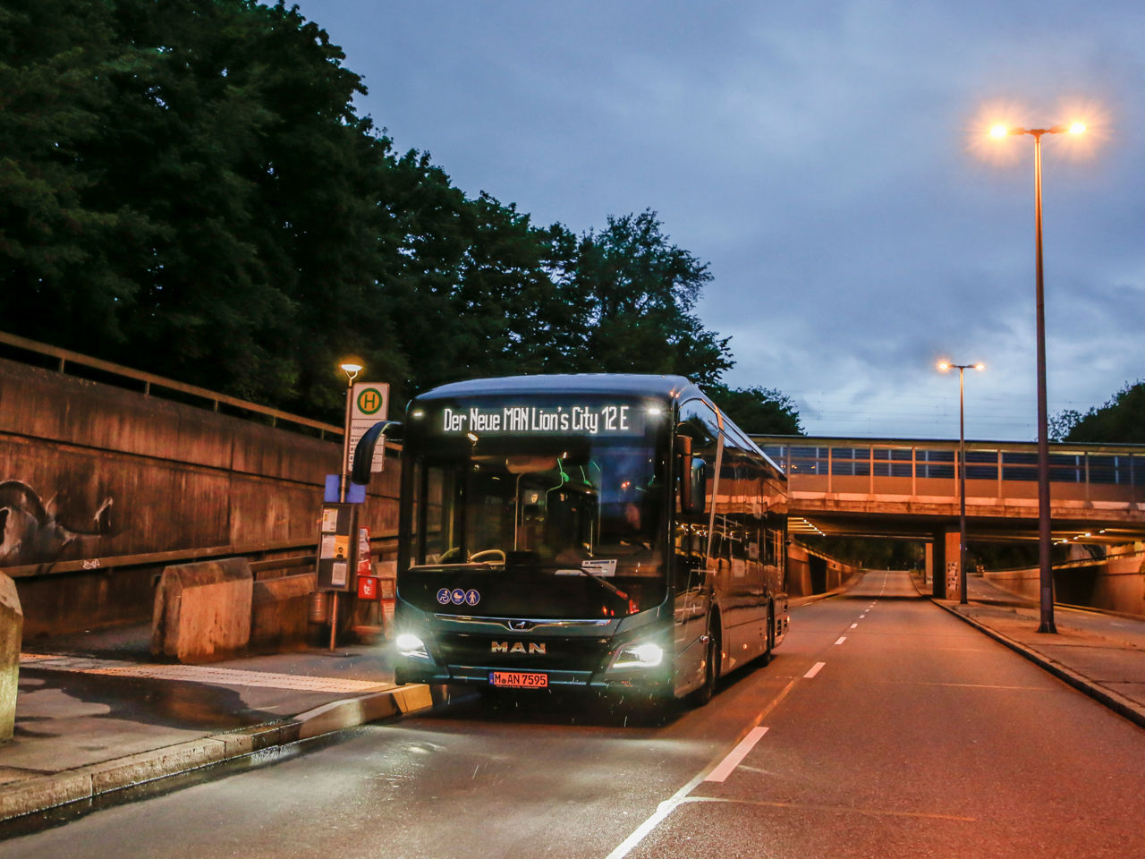 Ein MAN Lion's City 12 E steht in der Abenddämmerung an einer Bushaltestelle.