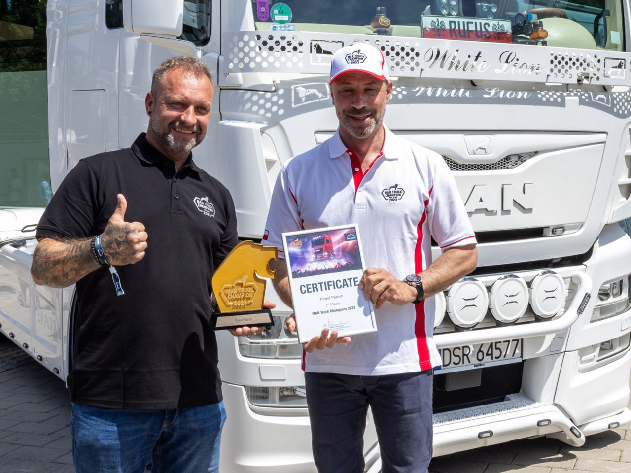 Two men stand in front of a white truck holding a certificate and a trophy