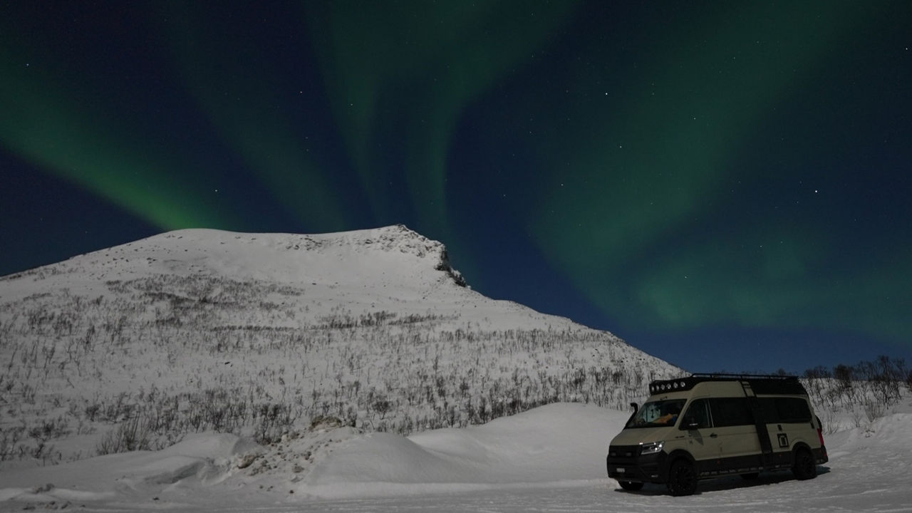 Kai Borstendorfer ist mit seinem MAN TGE im Norden von Europa unterwegs. Im Hintergrund sieht schneebedecke Berge und viele Polarlichter.