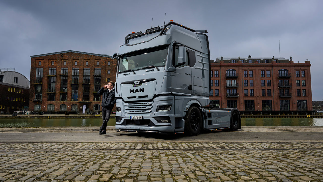 Ein grauer MAN TGX 18.560 mit D30-Motor steht vor Backsteingebäuden und sein Besitzer Tino Rateike lehnt am Lkw.