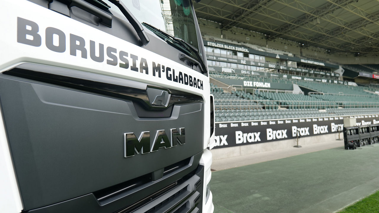 Der Kühlergrill des weißer MAN TGX mit Borussia Mönchengladbach Beklebung im Vordergrund und im Stadion von Borussia Mönchengladbach. Im Hintergrund sieht man die Ränge und Sitzplätze.