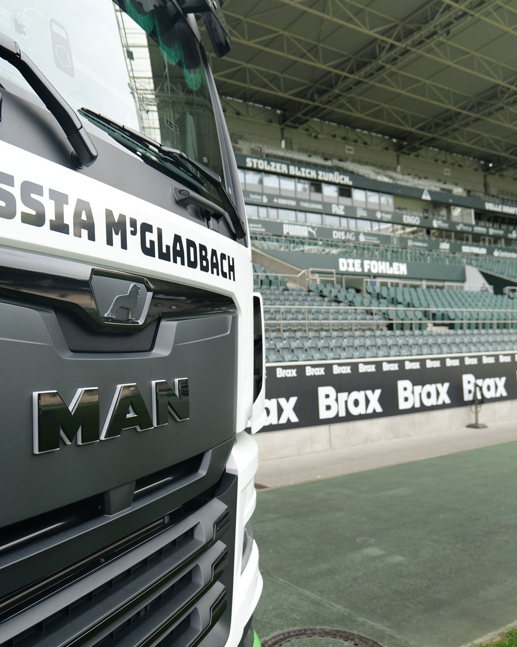 Der Kühlergrill des weißer MAN TGX mit Borussia Mönchengladbach Beklebung im Vordergrund und im Stadion von Borussia Mönchengladbach. Im Hintergrund sieht man die Ränge und Sitzplätze.