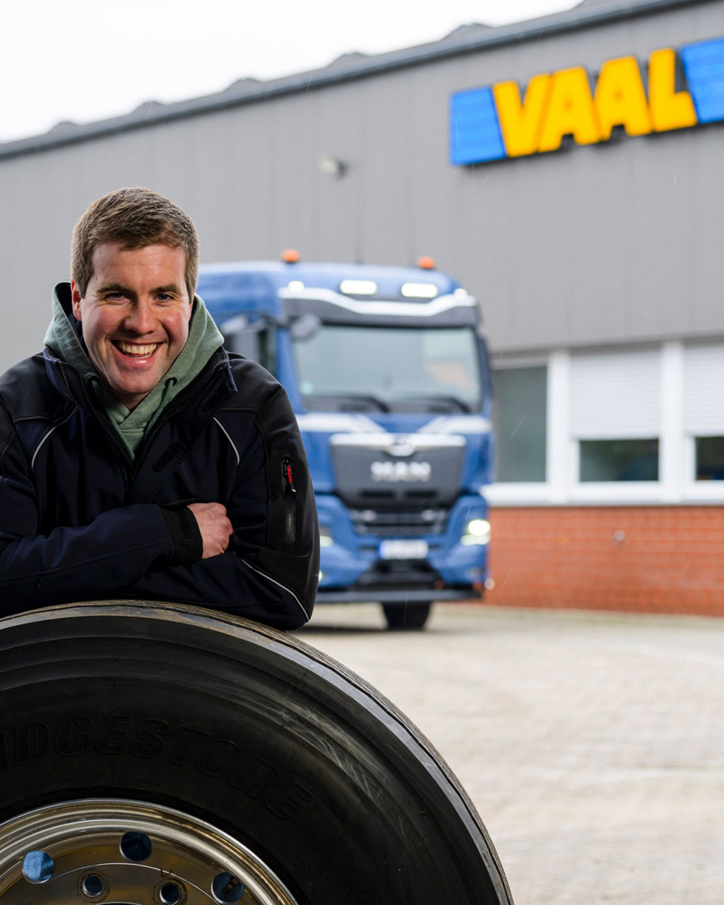 Ein Mitarbeiter von Vaal lehnt auf einem Reifen und im Hintergrund ist das Firmenlogo und der MAN eTruck zu sehen.