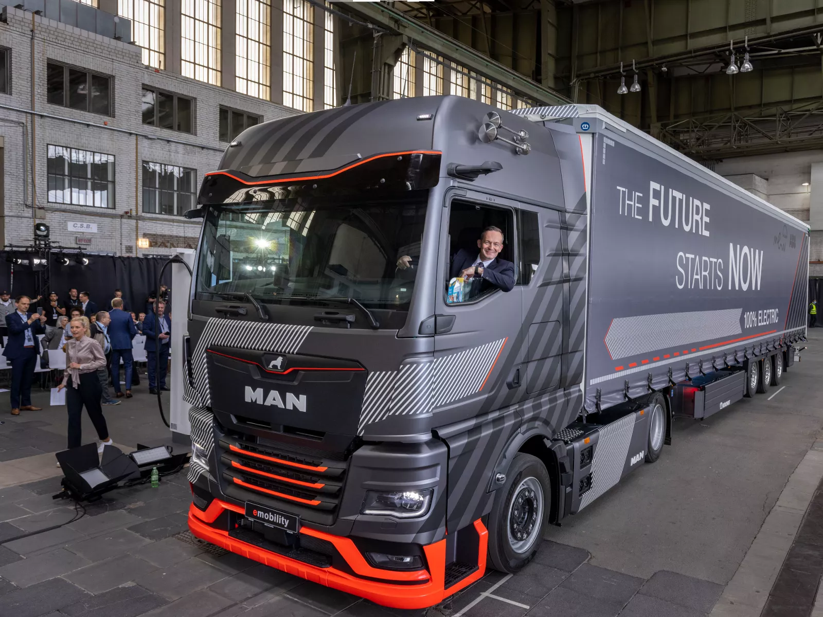 Driver inside MAN eTruck at indoor event, audience in background