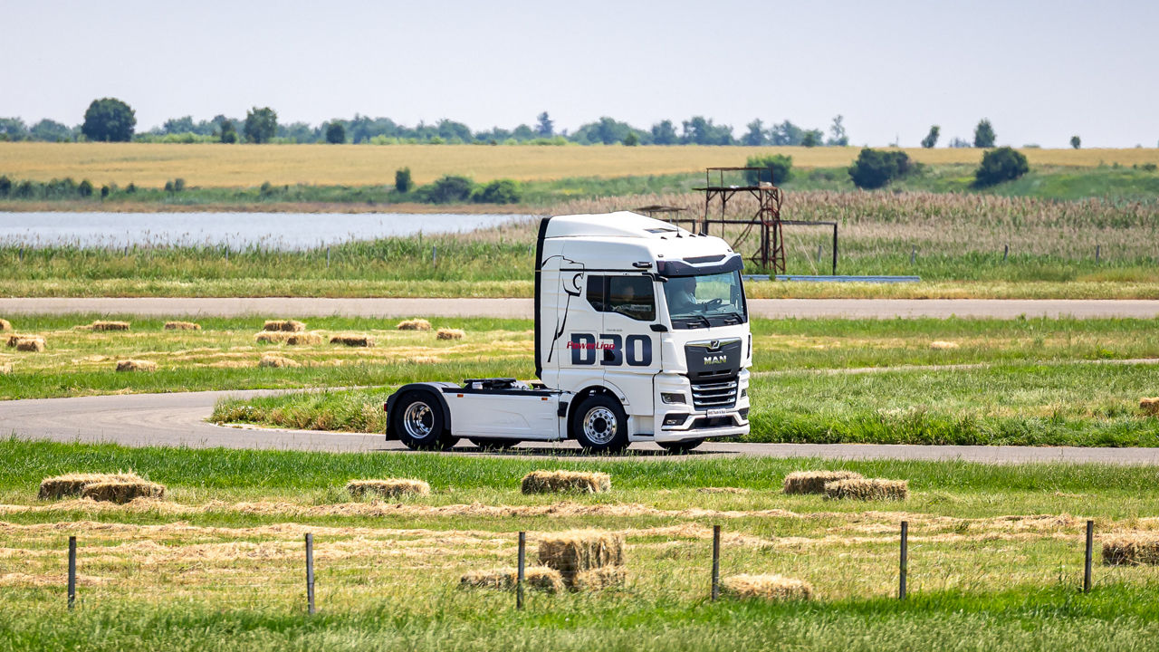 Ein weißer MAN TGX mit der Aufschrift „D30 PowerLion“ fährt auf einer Landstraße durch eine grüne Landschaft mit Heuballen und einem See im Hintergrund.