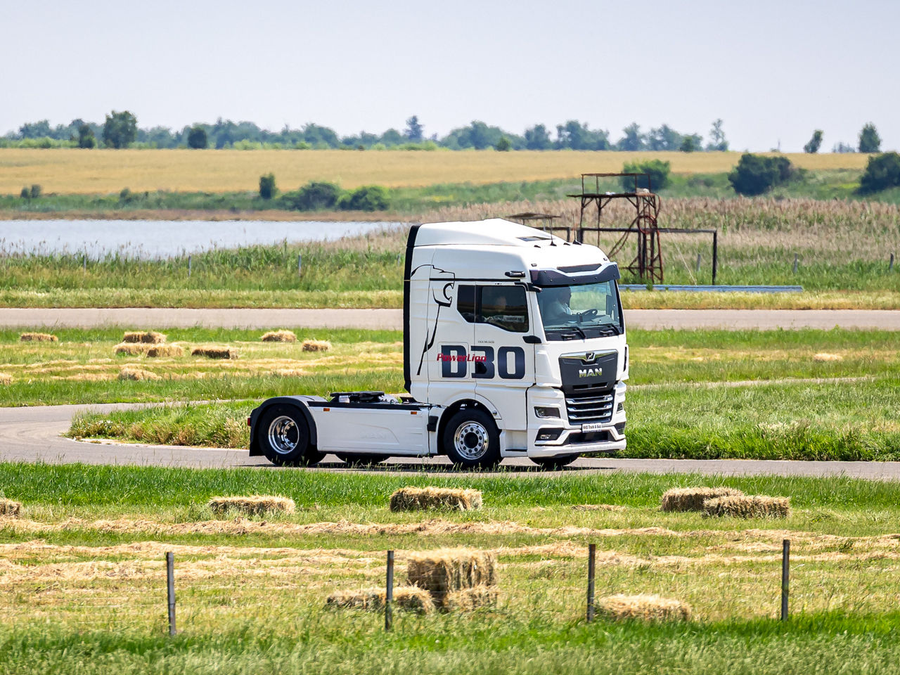 Білий MAN TGX з написом D30 PowerLion їде сільською дорогою серед зеленої місцевості на тлі тюків сіна та озера.