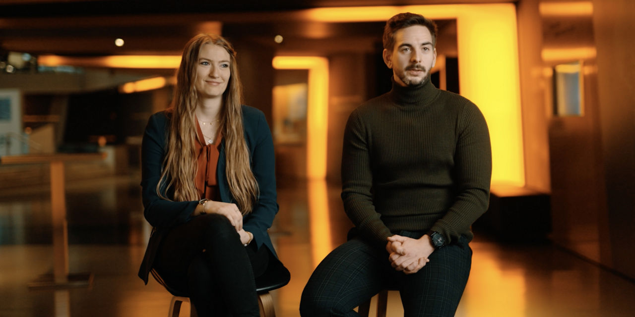 A woman and a man sit on chairs an look into the camera