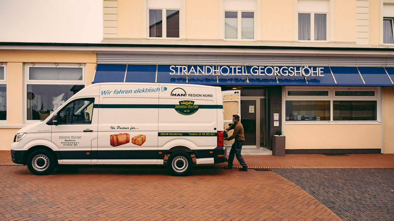 Ein MAN Speditionstransporter parkt vor einem Strandhotel. Eine Person hebt einen Koffer aus dem Wagen. 