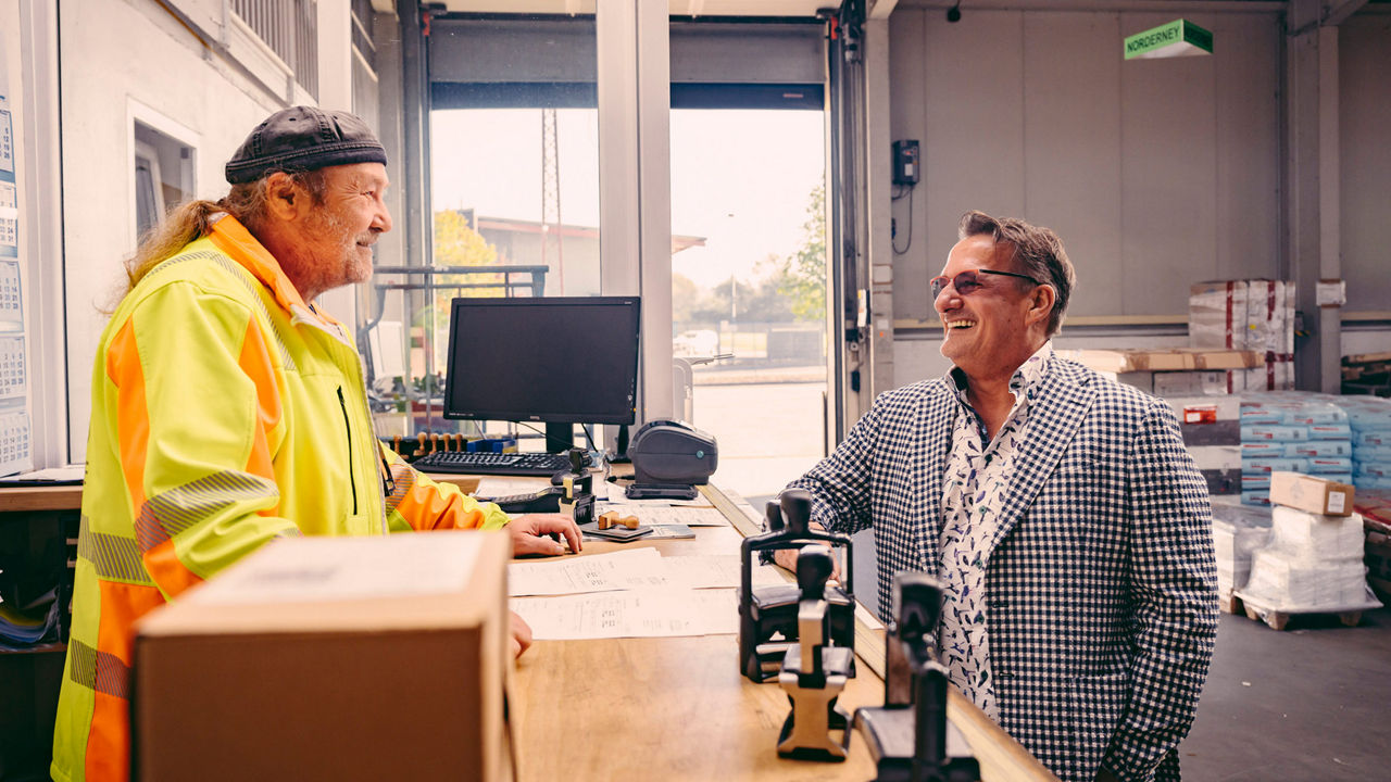 Spediteur Fischer steht an einem Tresen und unterhält sich mit einem Mann in Arbeitskleidung. 