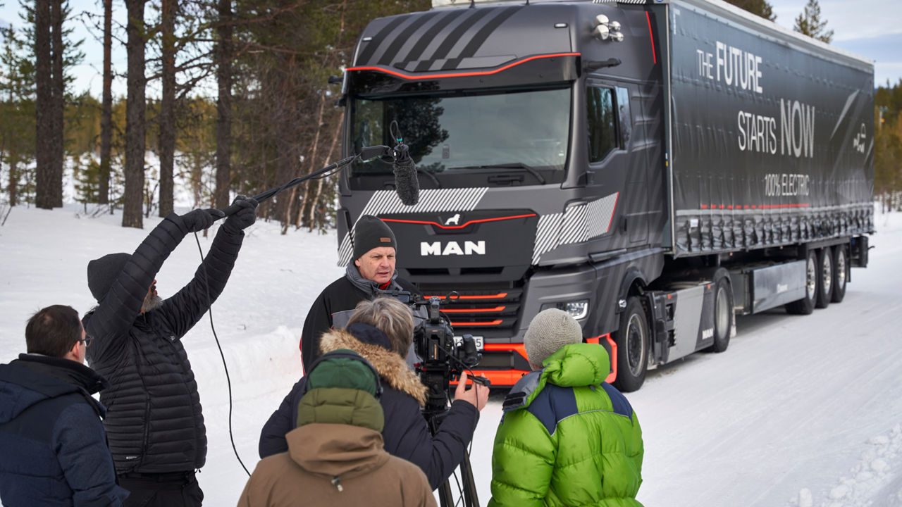 Rainer Miksch is standing in front of a MAN truck with several people and is being interviewed. In the background a winter landscape. 