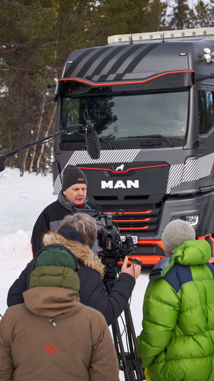 Rainer Miksch is standing in front of a MAN truck with several people and is being interviewed. In the background a winter landscape. 