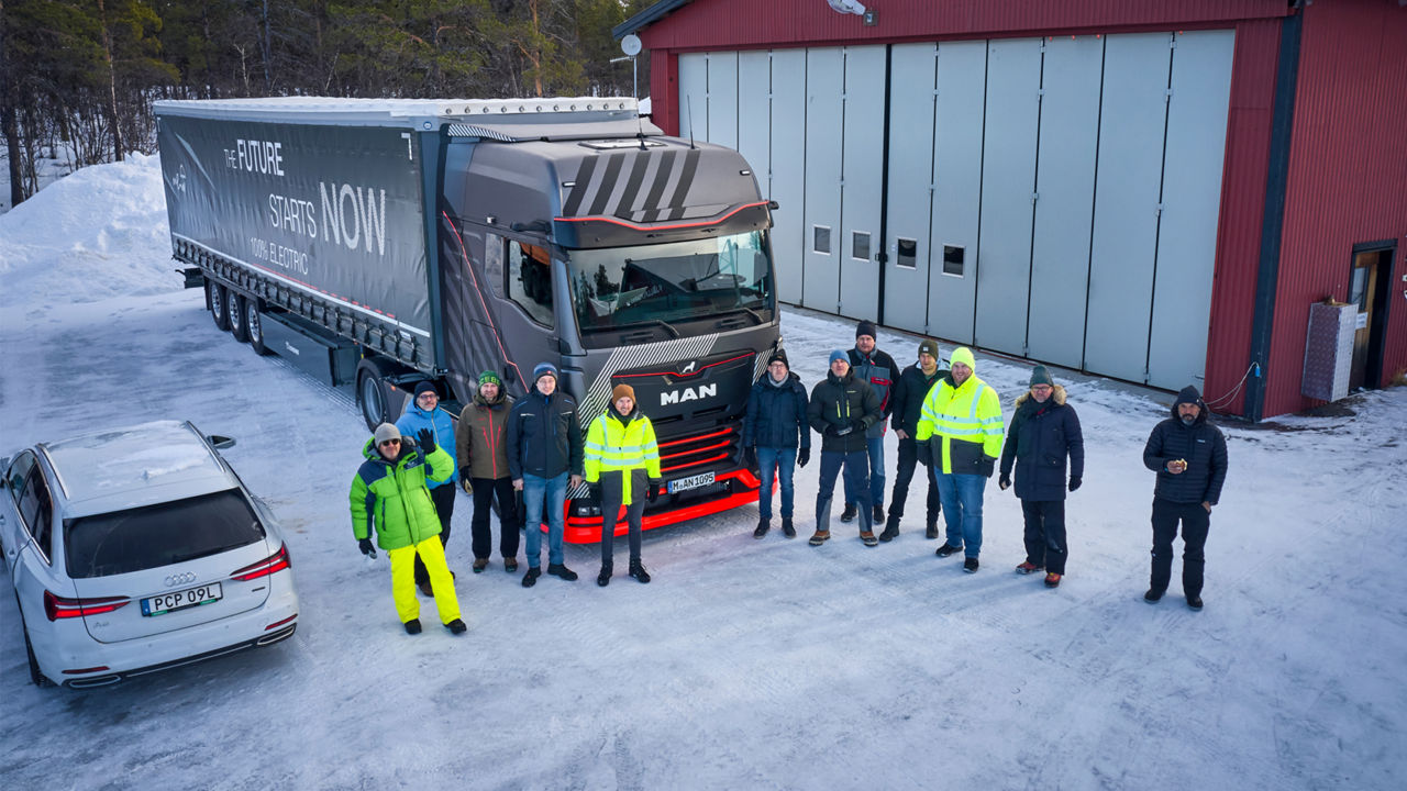 A group of people is standing in front of a MAN truck and a hall in a winter landscape. The people are looking at the camera. 