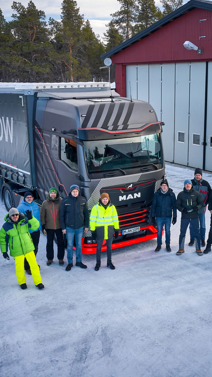 A group of people is standing in front of a MAN truck and a hall in a winter landscape. The people are looking at the camera. 