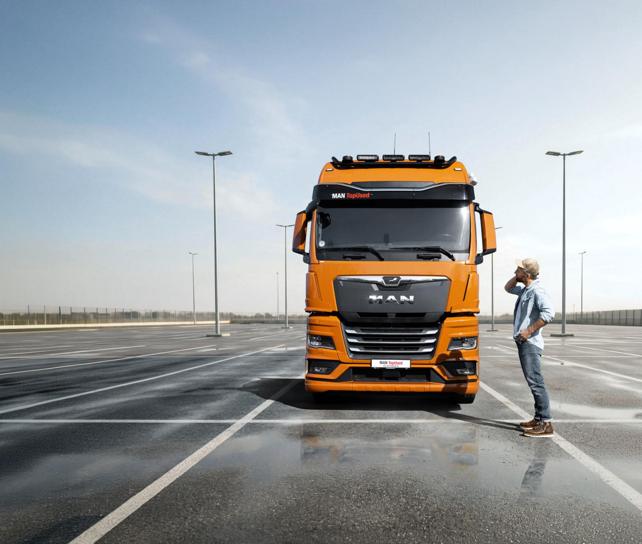 Camion arancione MAN TopUsed visto frontalmente in un ampio parcheggio bagnato, con un uomo in piedi accanto che osserva il camion per il trasporto su strada.
