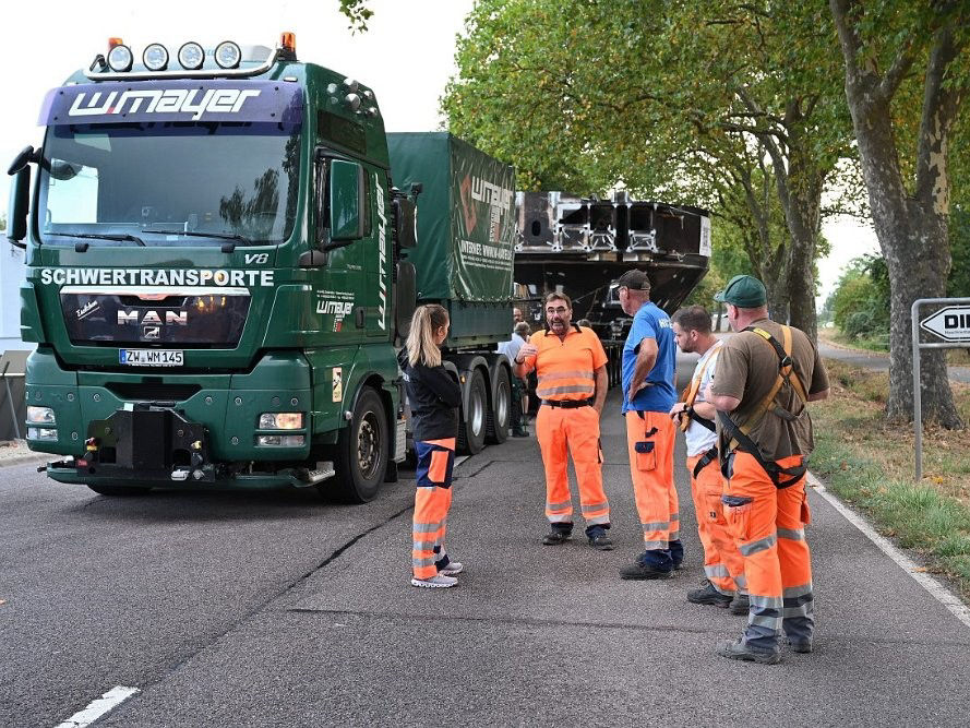 Ein MAN TGX der Firma W. Mayer mit Schwertransport steht am Straßenrand. Mehrere Arbeiter in orangefarbener Schutzkleidung besprechen sich vor dem Fahrzeug.