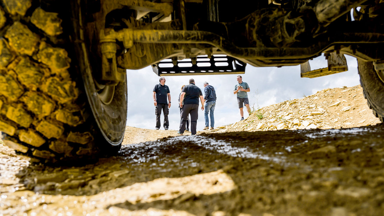 Mehrere Personen stehen auf einem unbefestigten, schlammigen Gelände und unterhalten sich. Das Bild ist aus der Perspektive unter einem großen, geländegängigen MAN Truck aufgenommen, dessen Reifen und Fahrwerk im Vordergrund sichtbar sind.
