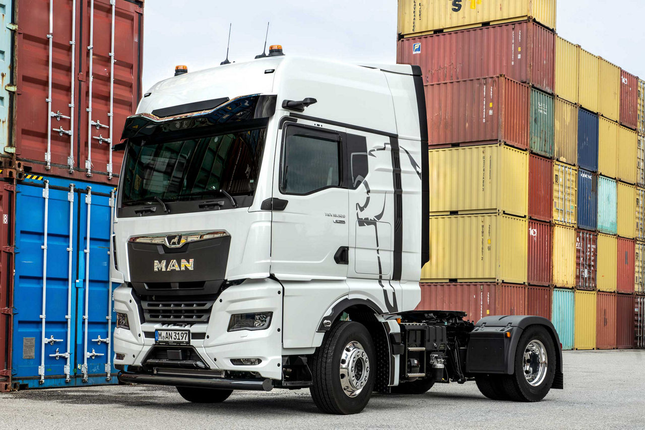 Ein weißer MAN-Lastwagen steht vor gestapelten bunten Frachtcontainern auf einem Hafengelände.